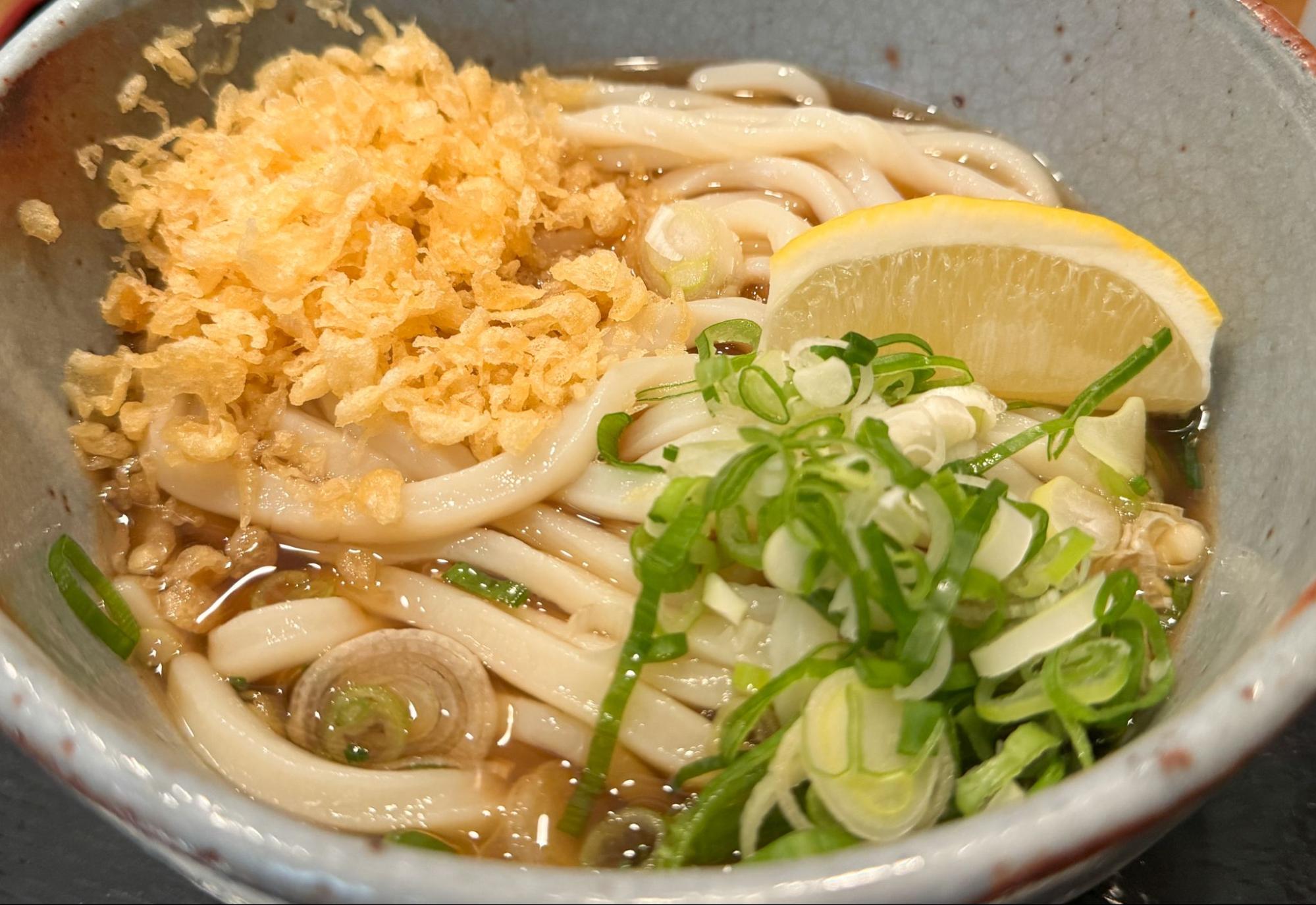 The udon is topped with green onions, tempura flakes, and lemon.